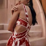 Woman wearing a red and white patchwork floral mini dress with side cutouts and wide straps, leaning against pastel stairs in a sunlit outdoor setting.

